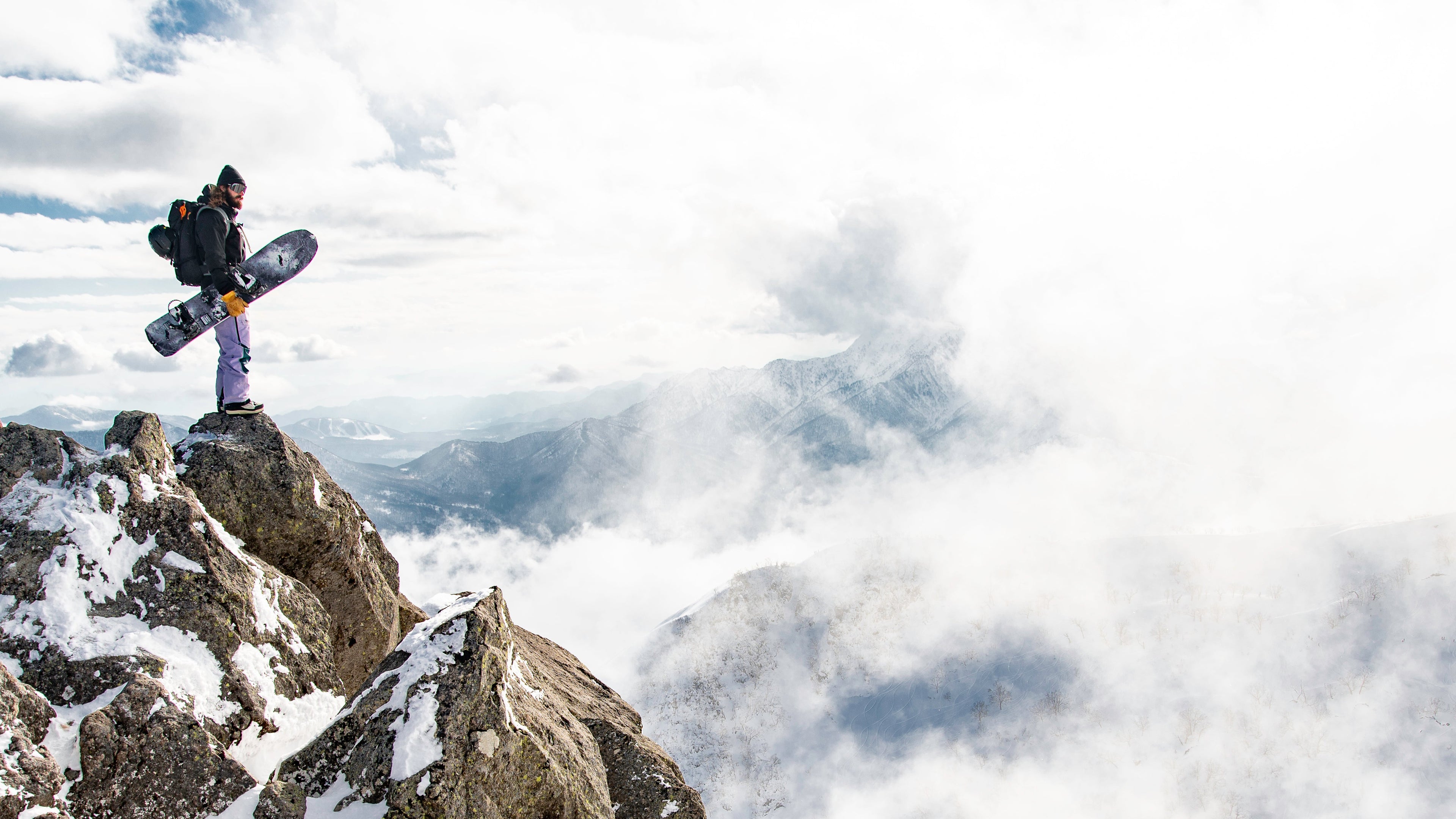 Person with a snowboard standing on a snowy mountain peak with clouds below